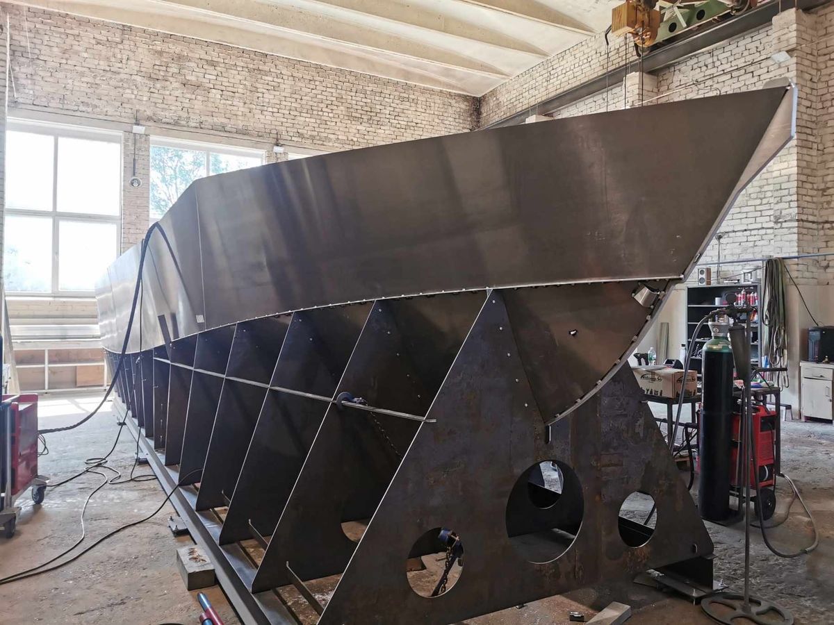 The AO12 hull under construction in a workshop, seen from the side on its jig with the transverse frames and cut-outs visible along the length