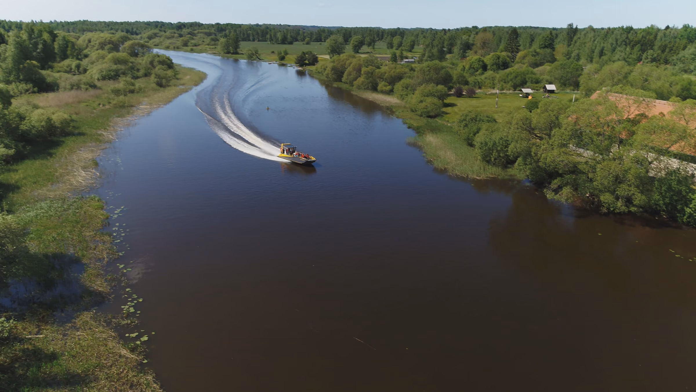 Aerial view of an LR9 Safari passenger boat running a narrow, winding river between wooded banks, passengers in lifejackets on the forward benches and a long clean wake curving behind