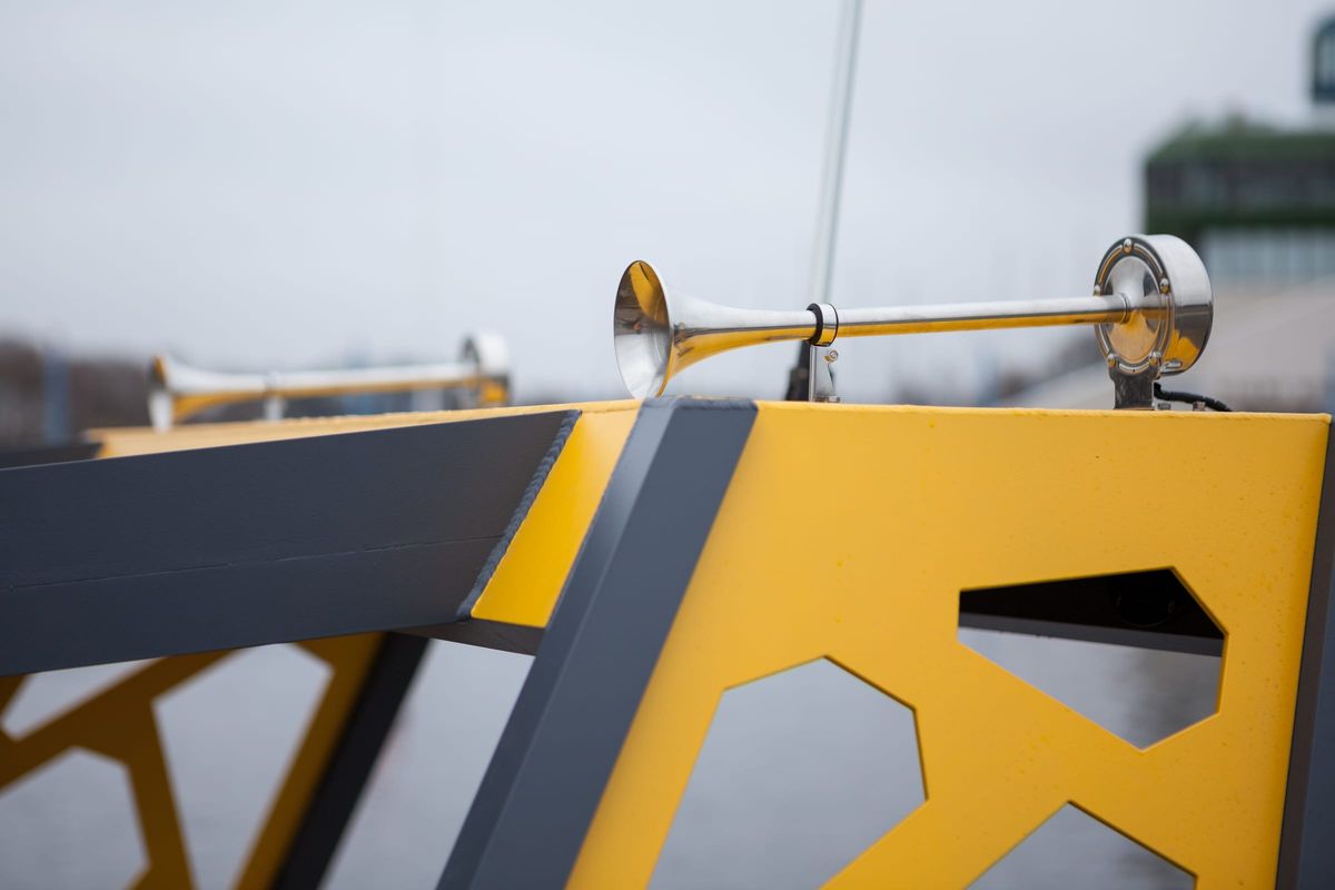 A chromed air horn mounted on the welded hardtop frame of the LR9 Safari passenger boat, the weld seam running along the beam and a cut-out yellow panel below it