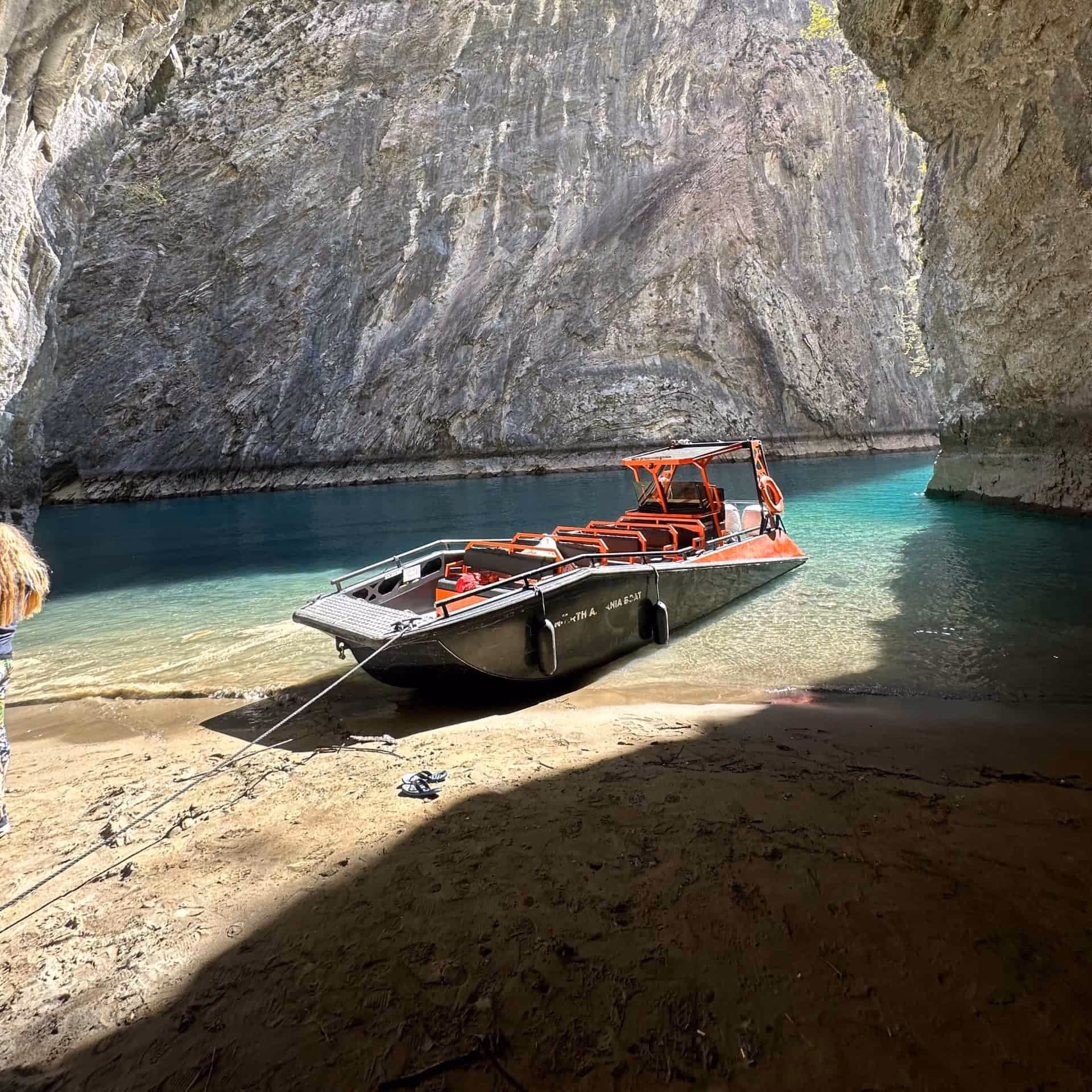 An LR9 Safari passenger boat beached on sand inside a limestone cave mouth, bow line run out to the beach, turquoise water behind it and a sunlit cliff wall rising above