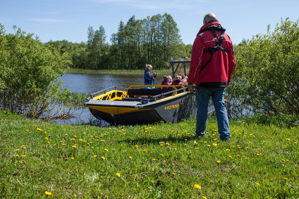 The LR9 Safari passenger boat nosed into a grassy riverbank with its bow platform lowered onto the grass, passengers still aboard and a crew member standing on the bank holding the bow