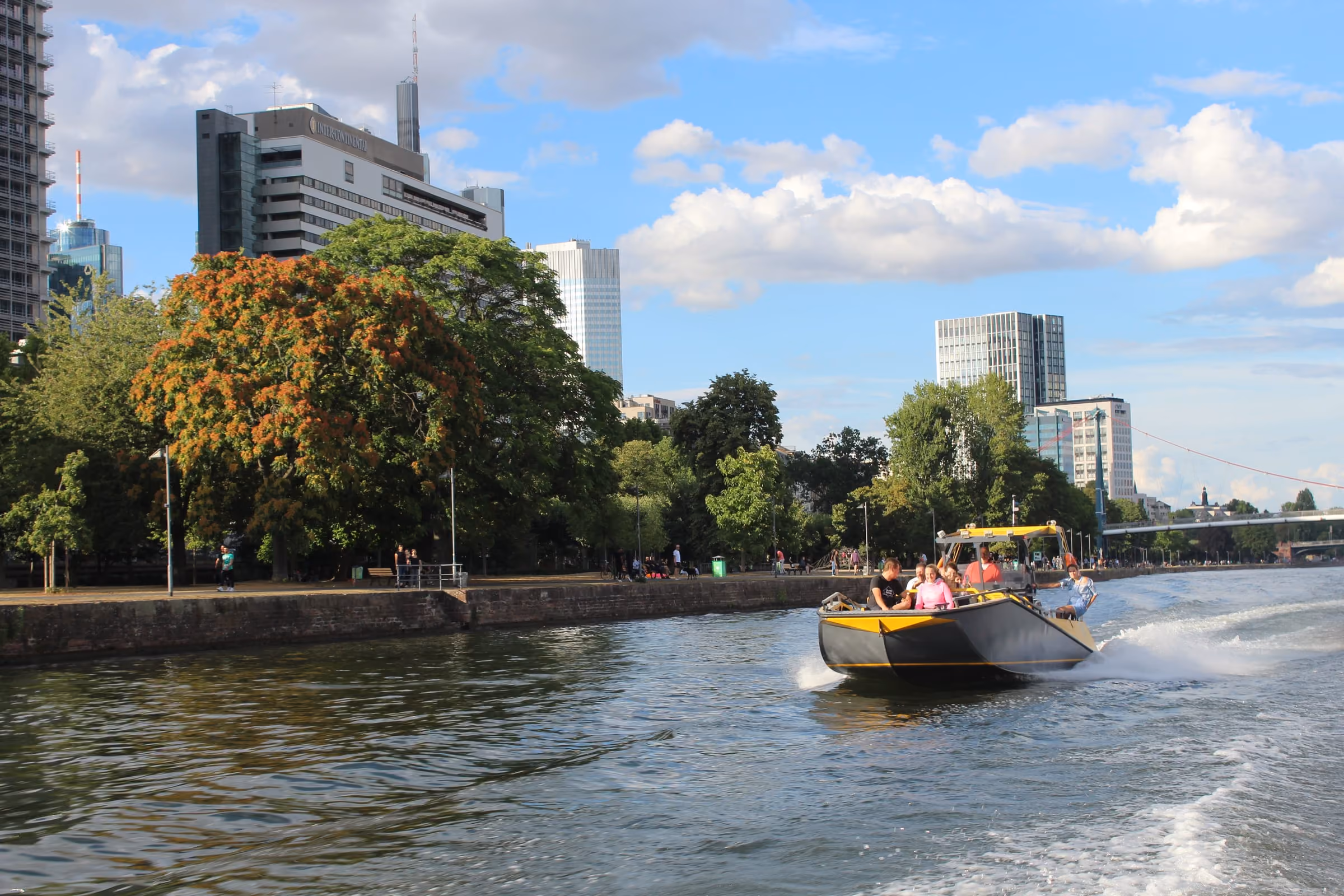 A black and yellow LR9 Safari passenger boat underway on the river Main in Frankfurt, passengers on the open forward deck and one crew member at the helm behind them, the city waterfront and high-rise skyline behind