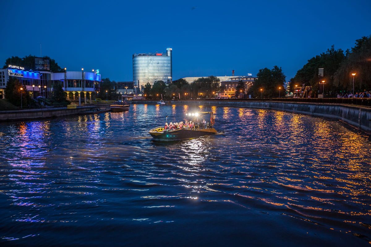 The LR9 Safari passenger boat underway on a city river at dusk with its deck floodlights lit and a full load of passengers aboard, lit buildings along the bank reflecting on the dark water