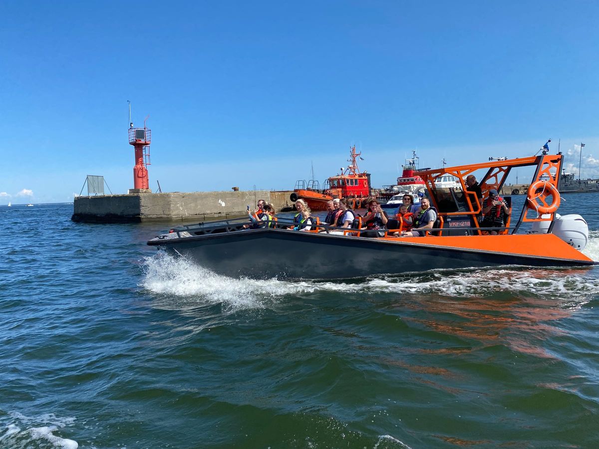 An orange LR9 Safari carrying passengers past a harbour breakwater and lighthouse, with tugs moored behind